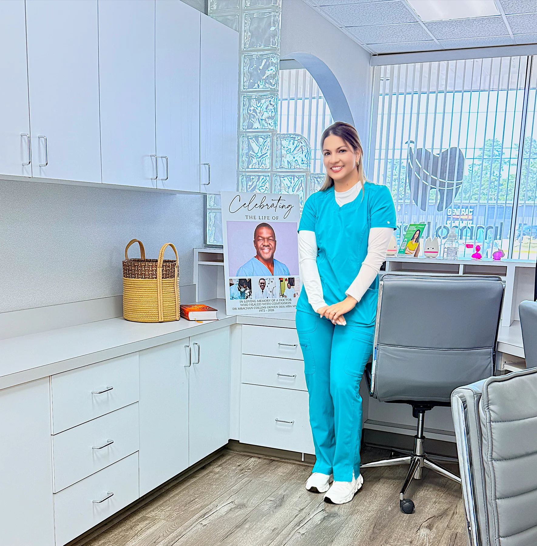 A team member in teal scrubs beside Dr. Okwen’s memorial portrait